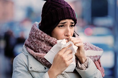 A woman is outside wearing a coat with a woolly hat and scarf holding a tissue to her face