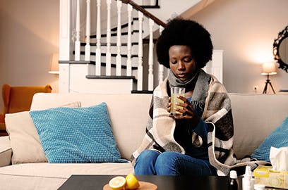 A woman is sitting on the sofa wrapped in a blanket drinking a drink from a glass