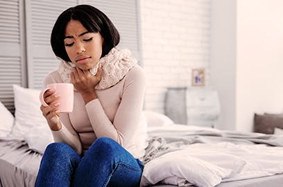 A woman is sitting on a bed with a scarf around her neck, holding a mug