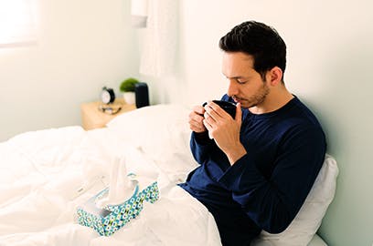 A man is sitting up in bed under a duvet drinking a hot drink from a mug