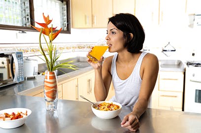 A woman is sitting at a kitchen counter top, she is drinking a glass of orange juice and there is a bowl of cereal in front of her