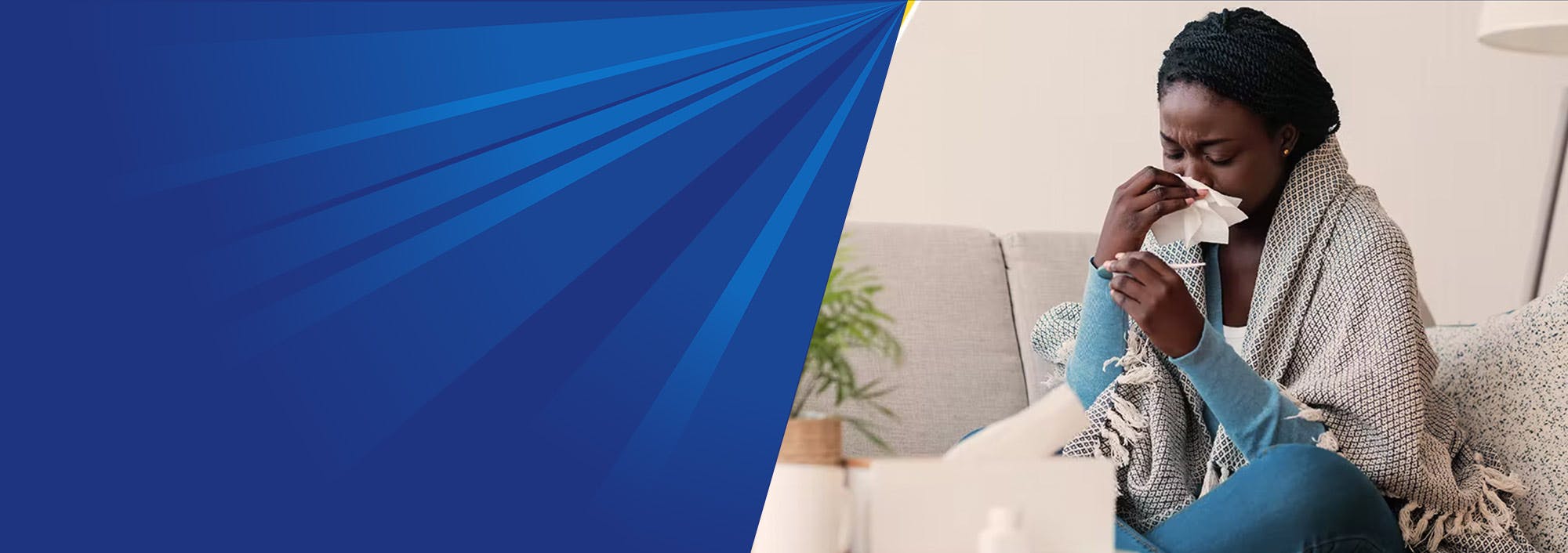 A woman is sat on a sofa. She is blowing her nose and looking at a digital thermometer