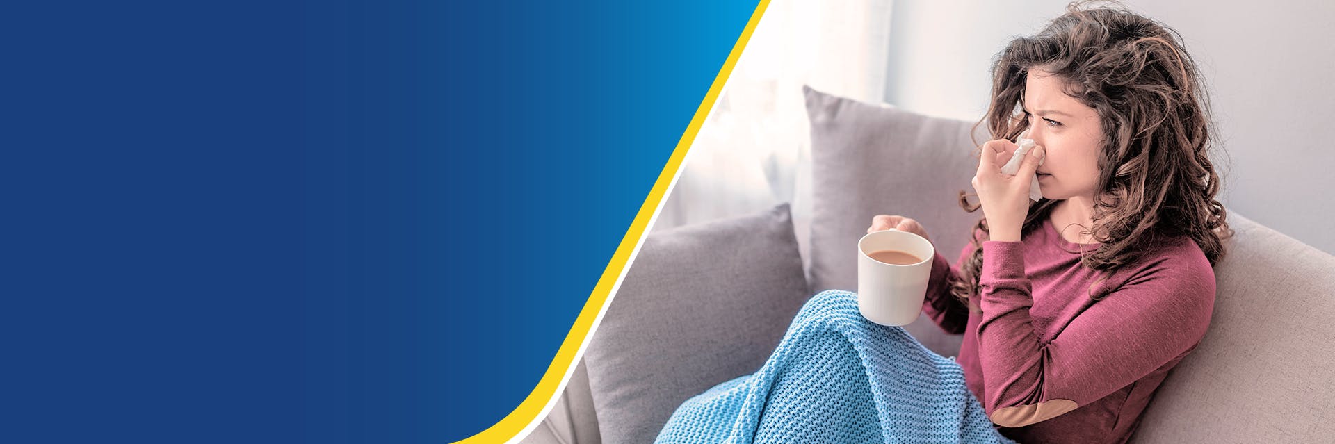 A woman is sitting on a sofa under a blanket holding a cup of tea and a tissue to her nose.  