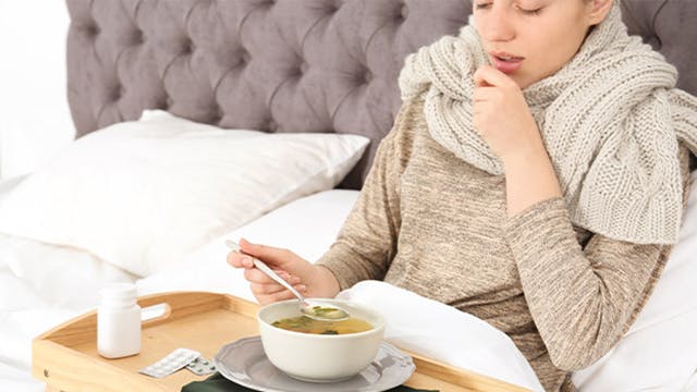 Young sick woman lying in bed eating broth off of a bed tray 