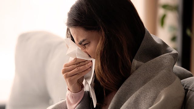 Woman sneezing into Kleenex with blanket around her shoulders 
