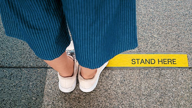 Person wearing blue pants and white shoes standing behind a yellow line on the sidewalk 