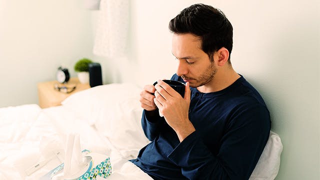 A man is sitting up in bed with a mug and a box of tissues