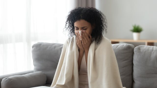 Young woman that is sick blowing her nose and huddling under a blanket. 