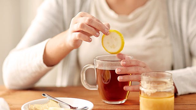 A woman is sitting at a table and adding a slice of lemon to a cup of tea. A plate of lemon and ginger slices and a pot of honey are also on the table.}