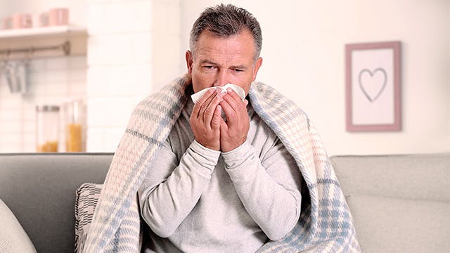 A man is sitting on the sofa under a blanket blowing his nose 