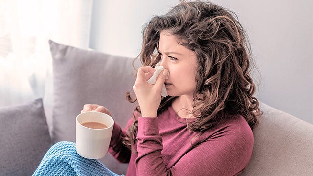 A woman is sitting on a sofa under a blanket holding a cup of tea and a tissue to her nose. 