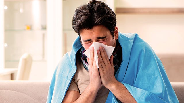 A man is sitting on the sofa wrapped in a blanket and holding a tissue to his face as if he is blowing his nose 