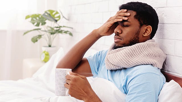 A man in bed is holding a mug and has a hand to his forehead 