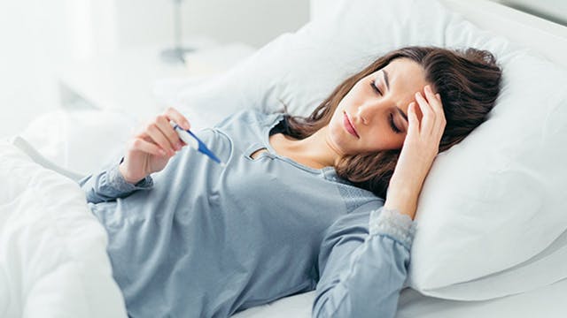 Young woman with flu lying in bed reading a thermometer 