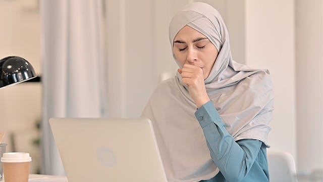 A woman sitting at a desk with a computer in front of her is coughing