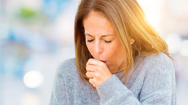 A woman in a gray sweater is outside coughing into fist