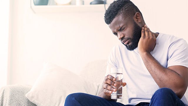 A man is seated on a sofa holding his neck. He looks in pain.