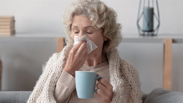 An older woman wrapped in a blanket is sitting on the sofa blowing her nose and holding a mug 