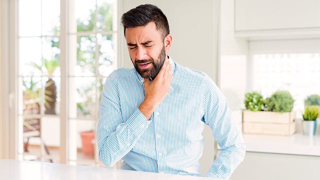 A man sat at a kitchen counter is holding his hand to his throat and looks in pain 