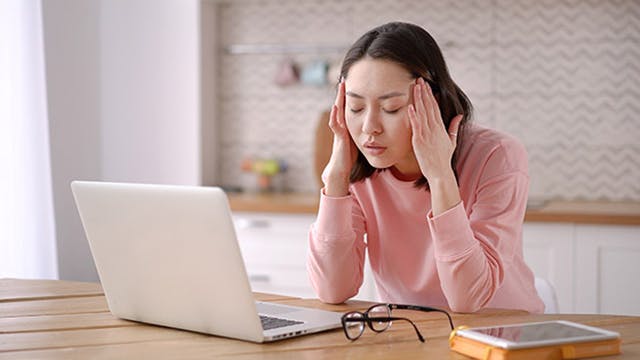 A woman who is working on a computer is holding her hands to her head in pain 