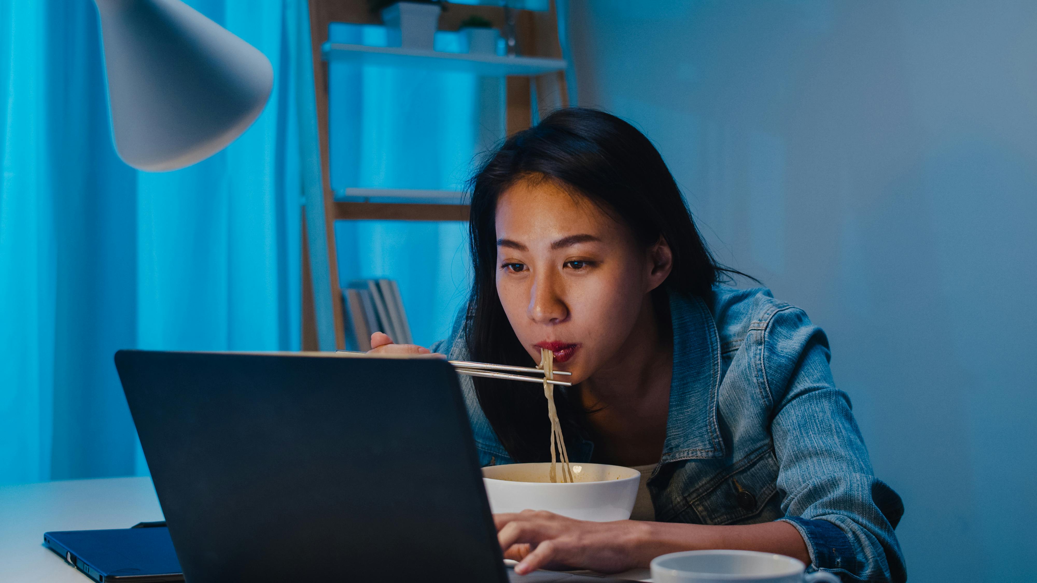Woman eating noodle soup in front of a laptop at night