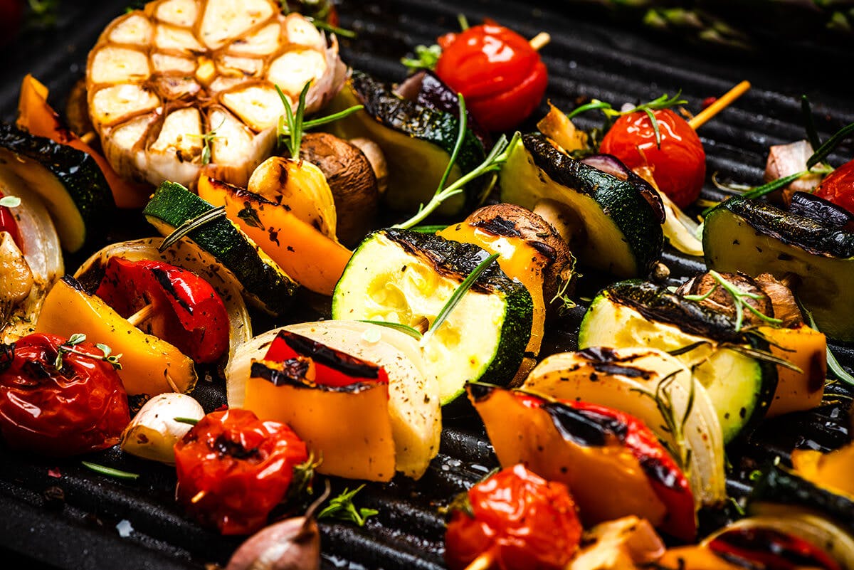 Three grilled vegetable skewers with tomatoes, squash, zucchini, garlic, and more on a barbecue grill