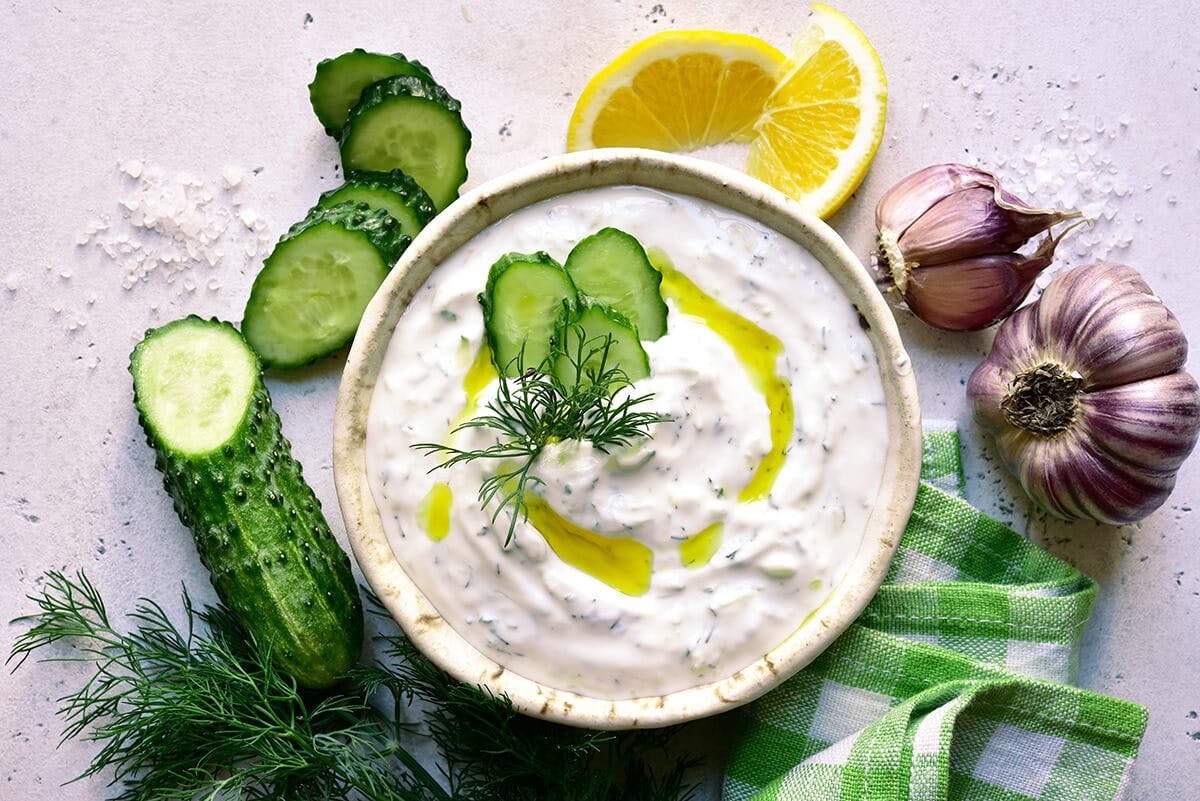 A bowl of Greek yogurt with dill, sliced lemons, cucumber, and purple garlic