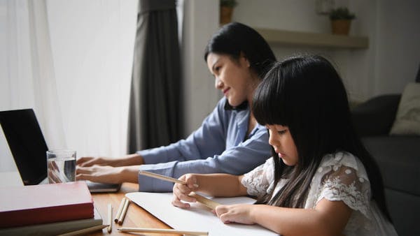 Mom working from home on laptop and phone while daughter draws next to her