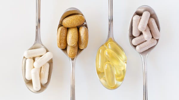 four spoons holding fish oil capsules, vitamins, and supplements with white background