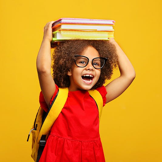 A smiling child with a missing front tooth wears a red dress, glasses and a yellow backpack while holding a stack of schoolbooks on her head