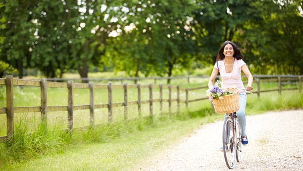 mujer joven montando una bicicleta cargando flores en la canasta delantera mientras está en un camino rural