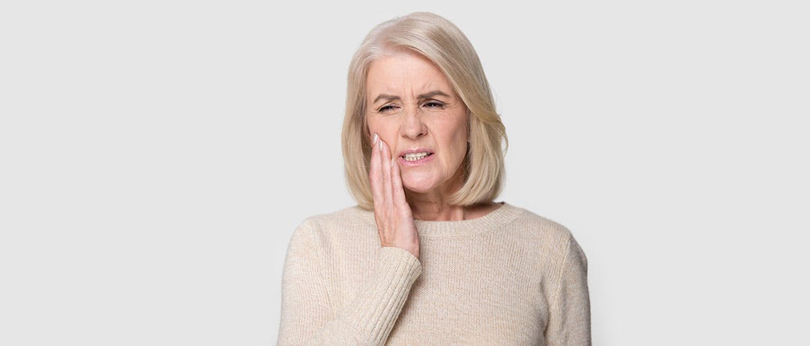 A blonde, elegant older woman in a beige jumper holds her hand up to her face as she winces with gum pain.
