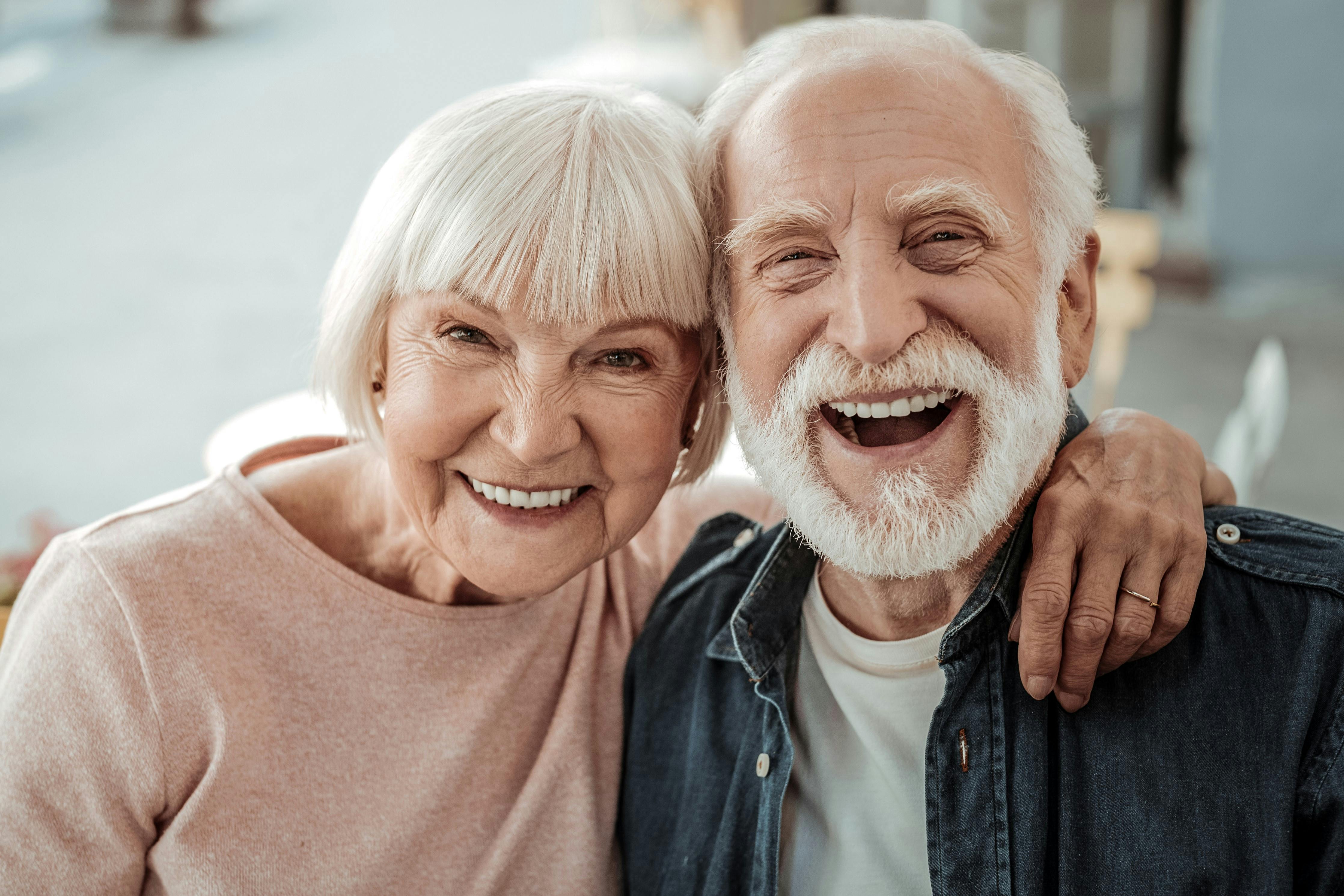 An attractive, elderly heterosexual couple lean their heads together as they smile joyfully into the camera.
