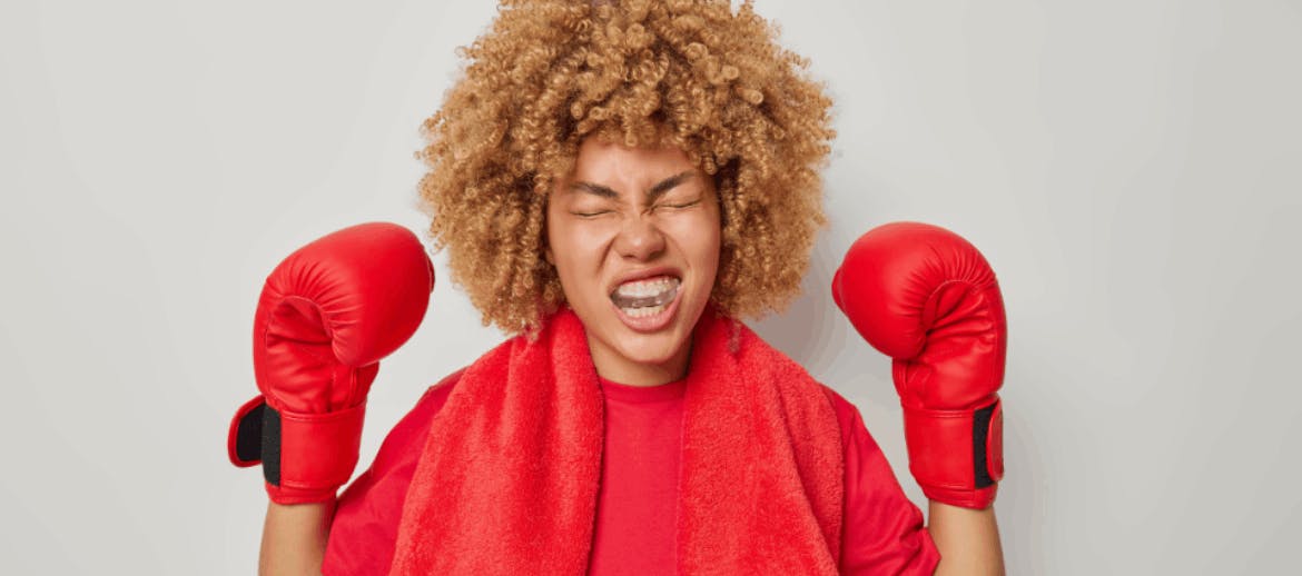 Energized female boxer with curly hair and raised arms wearing boxing gloves and protective mouth guard