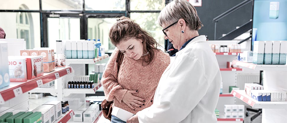 Eine Patientin, die an Schmerzen leidet, im Gespräch mit einem Apotheker; beide schauen sich gemeinsam ein Präparat an.