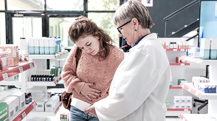 Eine Patientin, die an Schmerzen leidet, im Gespräch mit einem Apotheker; beide schauen sich gemeinsam ein Präparat an.