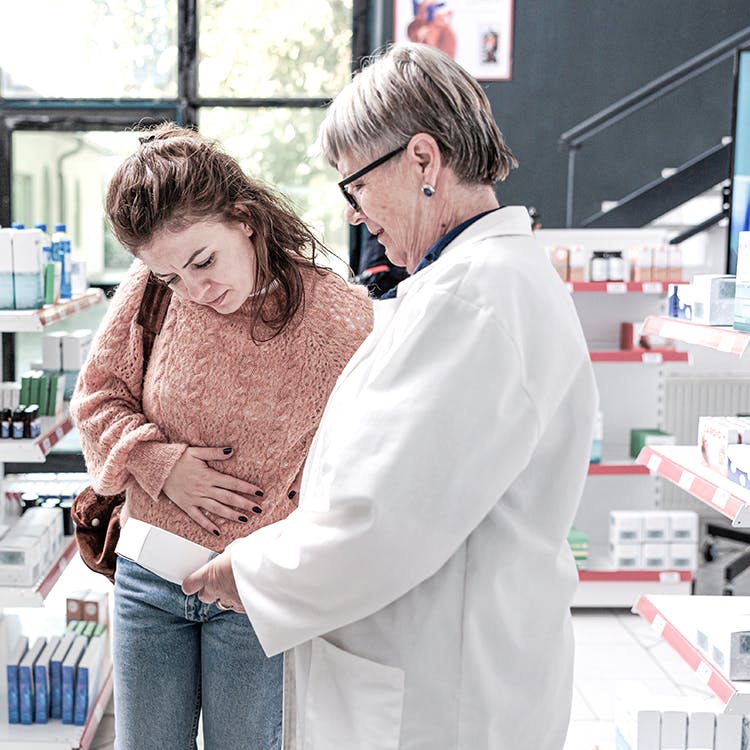 Eine Patientin, die an Schmerzen leidet, im Gespräch mit einem Apotheker; beide schauen sich gemeinsam ein Präparat an.