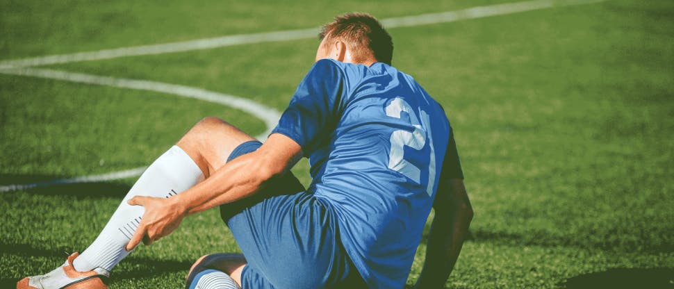 Mann mit kurzem hellbraunem Haar auf einem Fußballfeld, der ein Fußballtrikot trägt und vor Schmerzen sein Bein umklammert