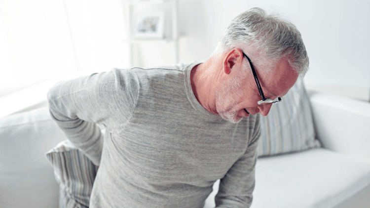 Ένας μεσήλικας με γκρίζα μαλλιά και γυαλιά αγγίζει τη μέση του και μορφάζει σαν να υποφέρει από πόνο στη μέση