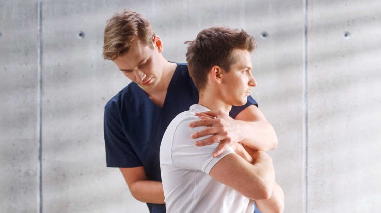 Ένας νεαρός ασθενής με λευκό μπλουζάκι υποβάλλεται σε θεραπεία για μυϊκά διαστρέμματα και καταπόνηση από έναν φυσιοθεραπευτή με μπλε στολή.