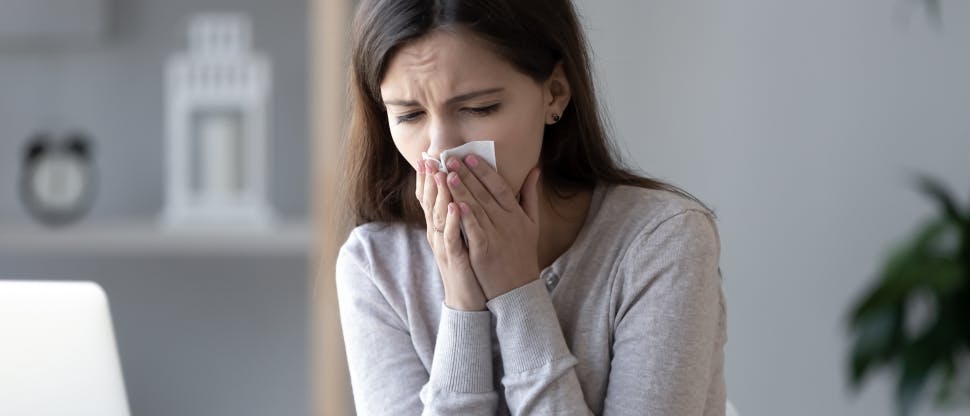 A young brunette woman sits at home and blows her nose, looking  as if she is in pain.