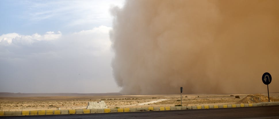 Image of a dust storm over a road and desert plain. The dust cloud takes over half  of the sky