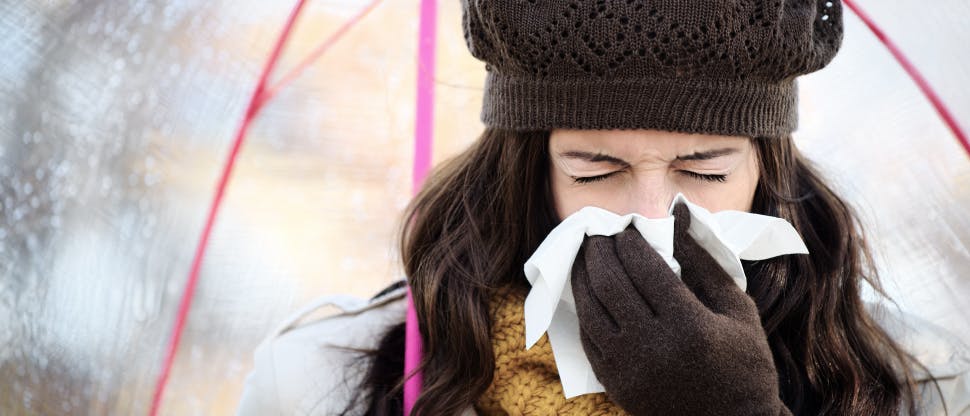 Young woman wearing a brown knitted hat and gloves blows her nose into a tissue while standing under an umbrella