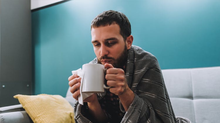 Young man sits on a grey sofa with a striped blanket draped  over his shoulders holding a mug of coffee and a tissue