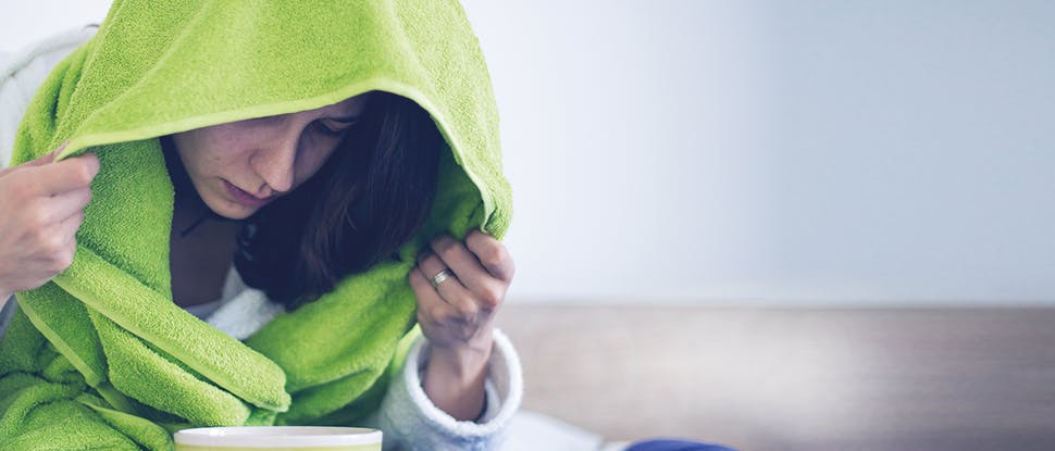 Young brunette woman holds a green towel over her head as she  inhales steam from a bowl of hot water to clear nasal congestion