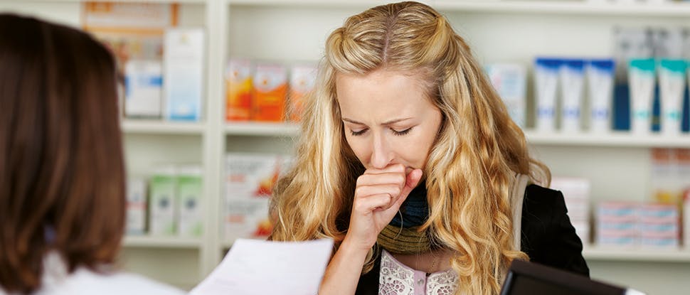 Young woman with long wavy blonde hair stands  in front of a female brunette pharmacist in a  pharmacy coughing