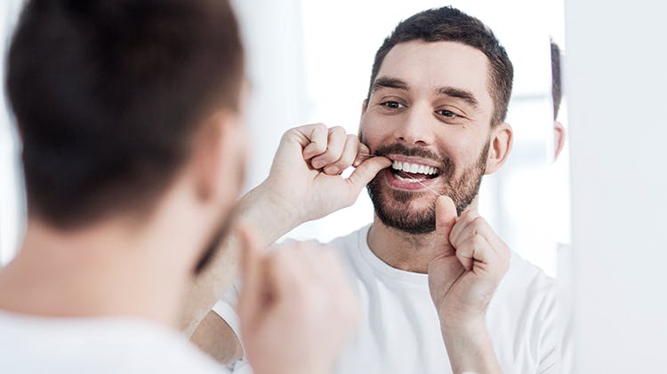 A man flosses his teeth in a bathroom mirror.