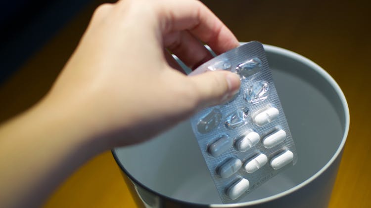 A hand places an expired, used pill packet in a safe disposal area to dispose of the medicine correctly.