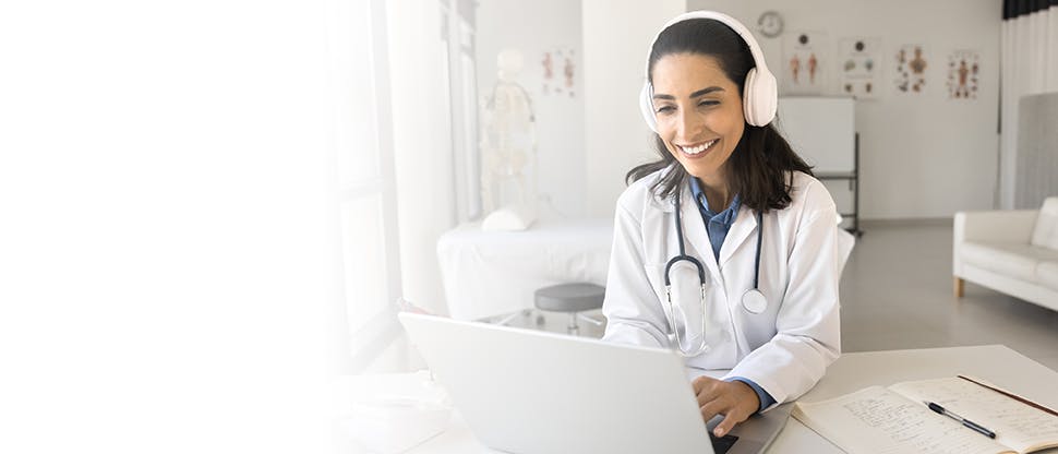 Woman doctor working at laptop in hospital office, typing, smiling, using wireless headphones for video conference call.