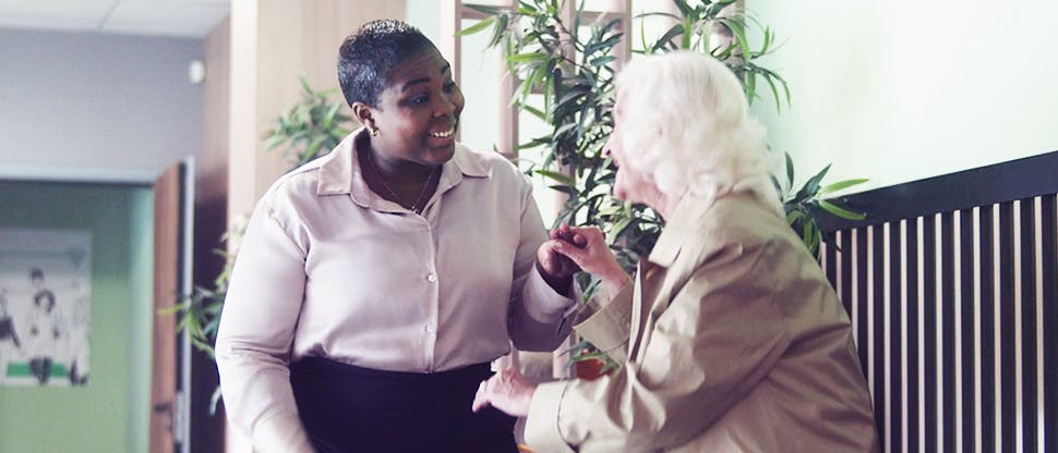 A GP helping an elderly lady up from her seat in the surgery waiting room